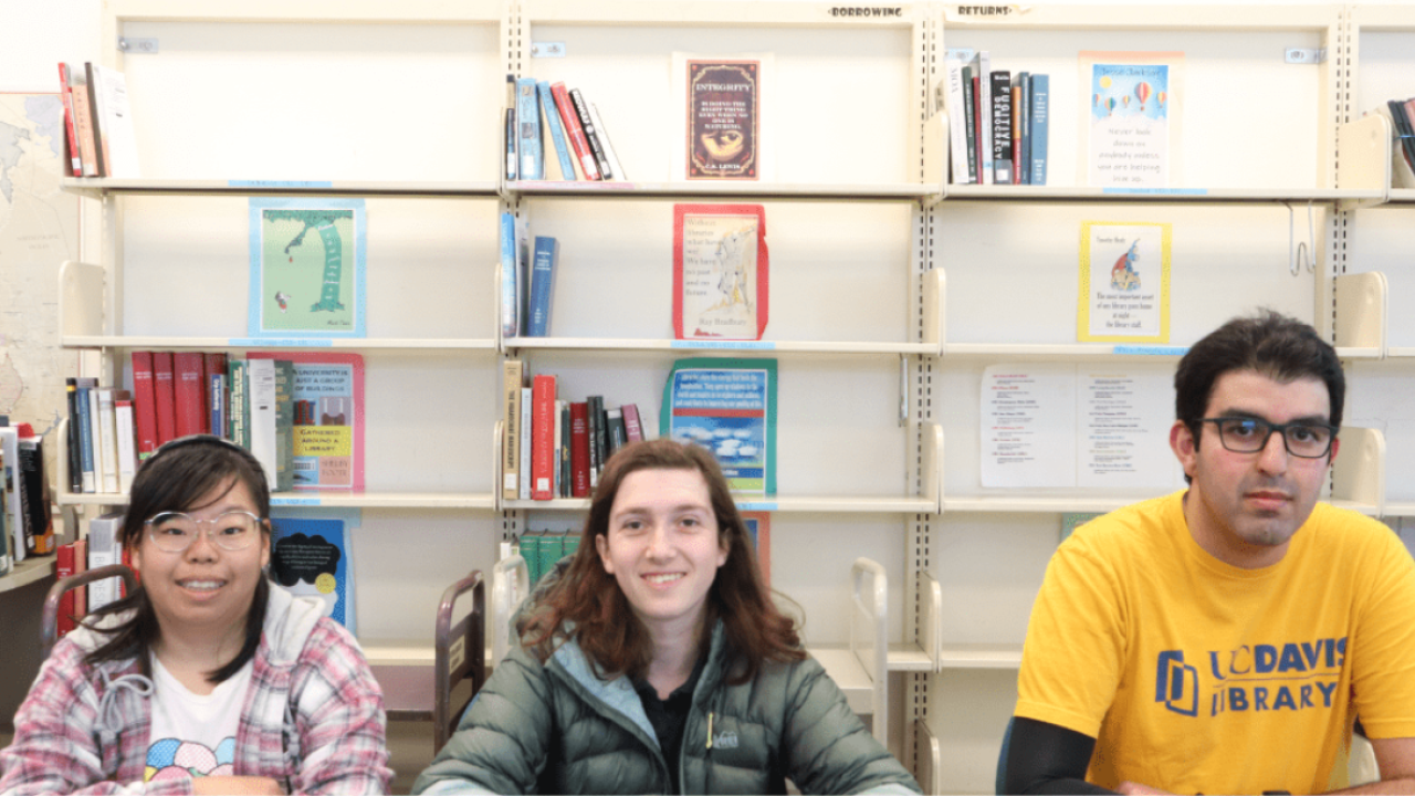 three students sitting at table with bookshelves behind them