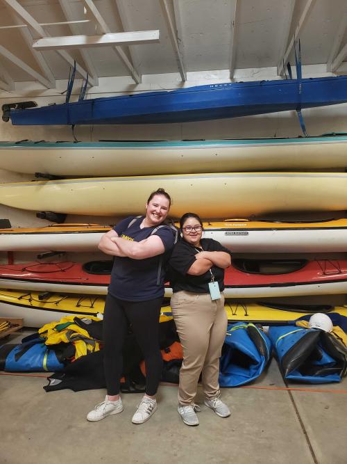 a student and mentor standing back to back with arms crossed in front of canoes